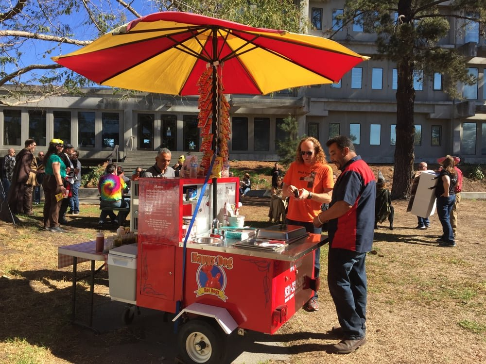 The Happy Dog Hotdogs red cart serving customers under a yellow and red umbrella at an outdoor event in Santa Cruz